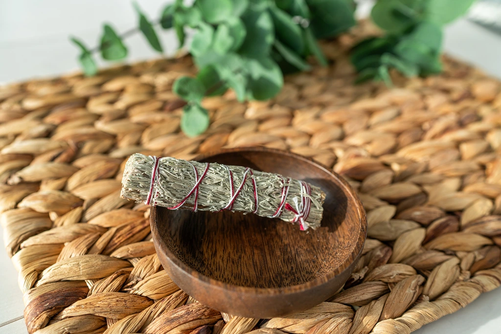 sage smudge sitck in a wooden bowl on top of a woven placement with leaves 