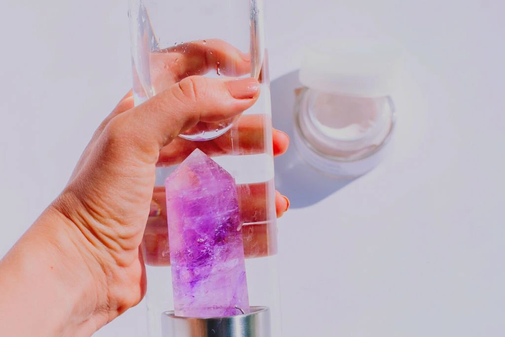 A person's hand holding a water bottle with an amethyst crystal inside it on a white background.