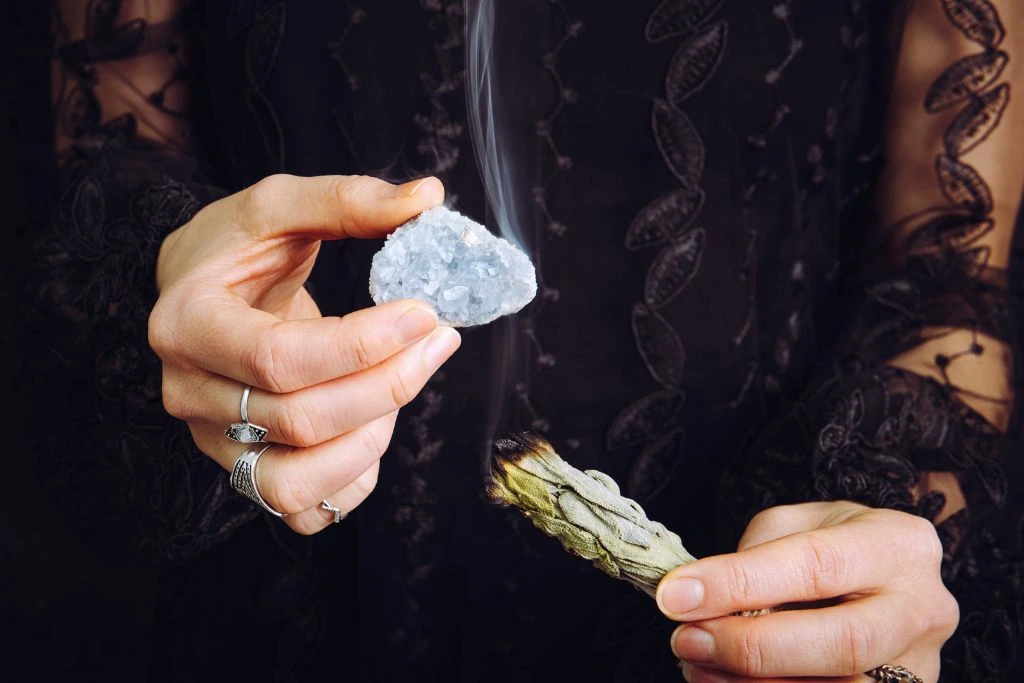 A person holding a celestite crystal above the other hand with a burning sage smudge stick