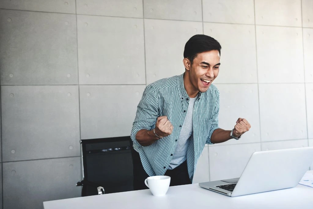 A man celebrating his success in front of his laptop