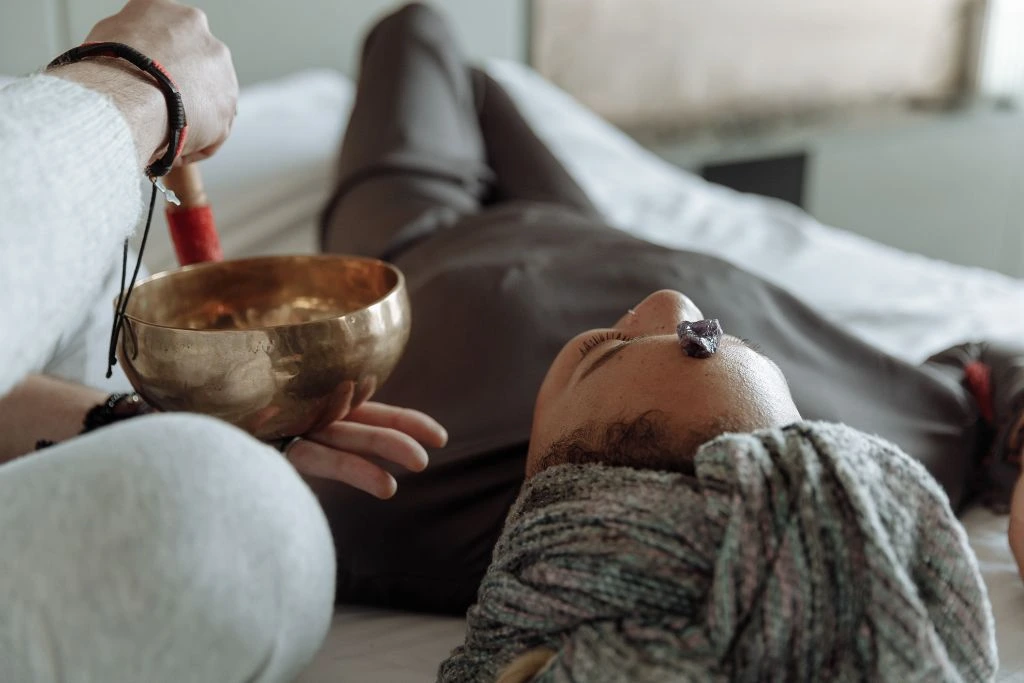 A person lying down with a crystal cluster on the forehead and another person using a tibetan singing bowl