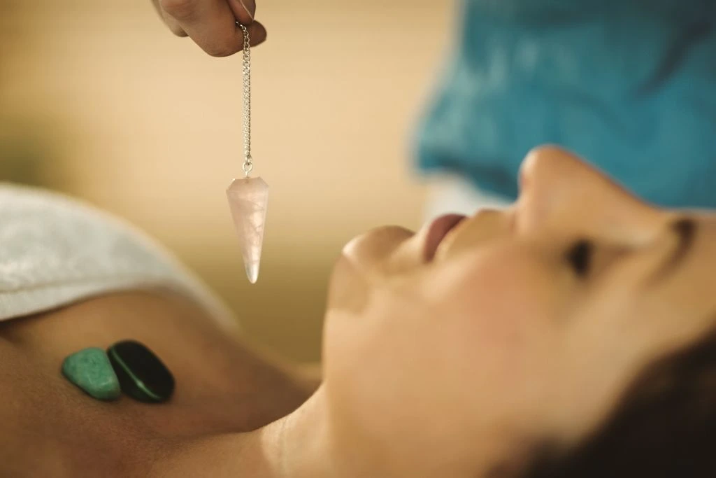 A woman with crystals on her chest while a person is holding a crystal pendulum above it