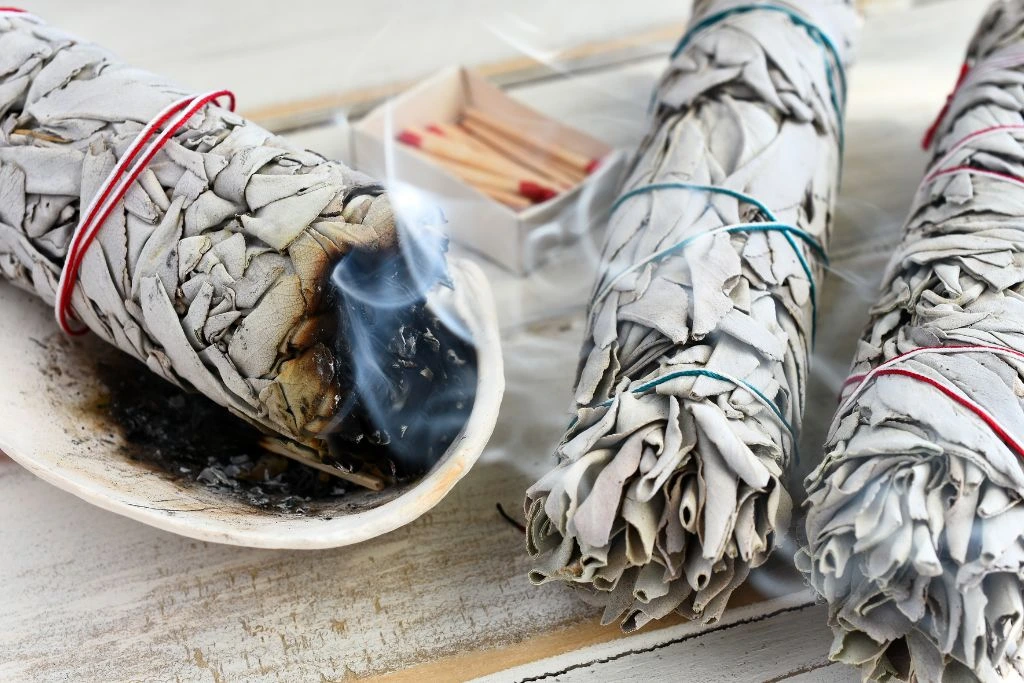 A burning sage smudge stick in the bowl with two other sage smudge sticks