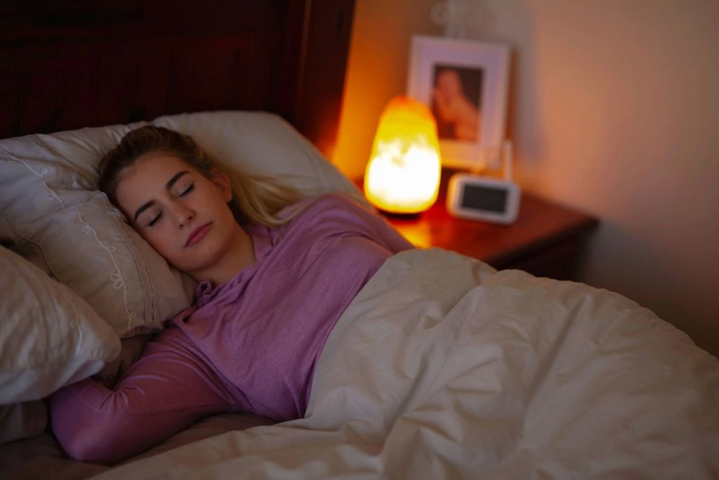 a woman sleeping on her bed while her Himalayan salt lamp is on placed on her bed side table