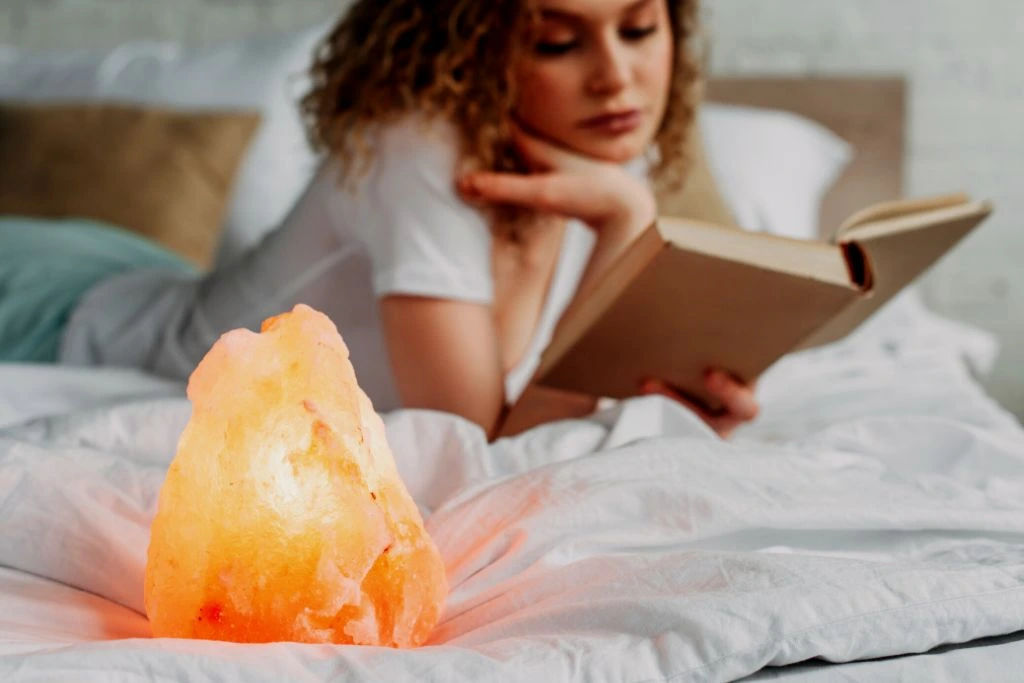 Himalayan salt lamp placed on bed with a blurry woman reading a book on the background