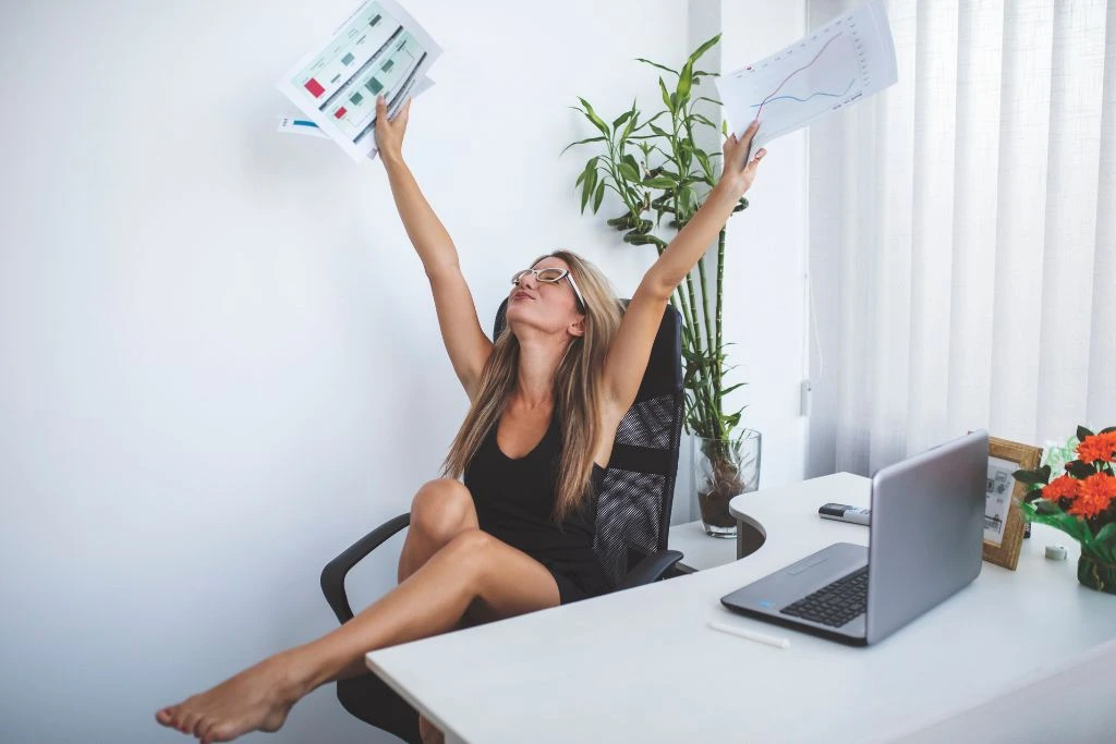 A woman raising her hands while holding papers in front of her laptop