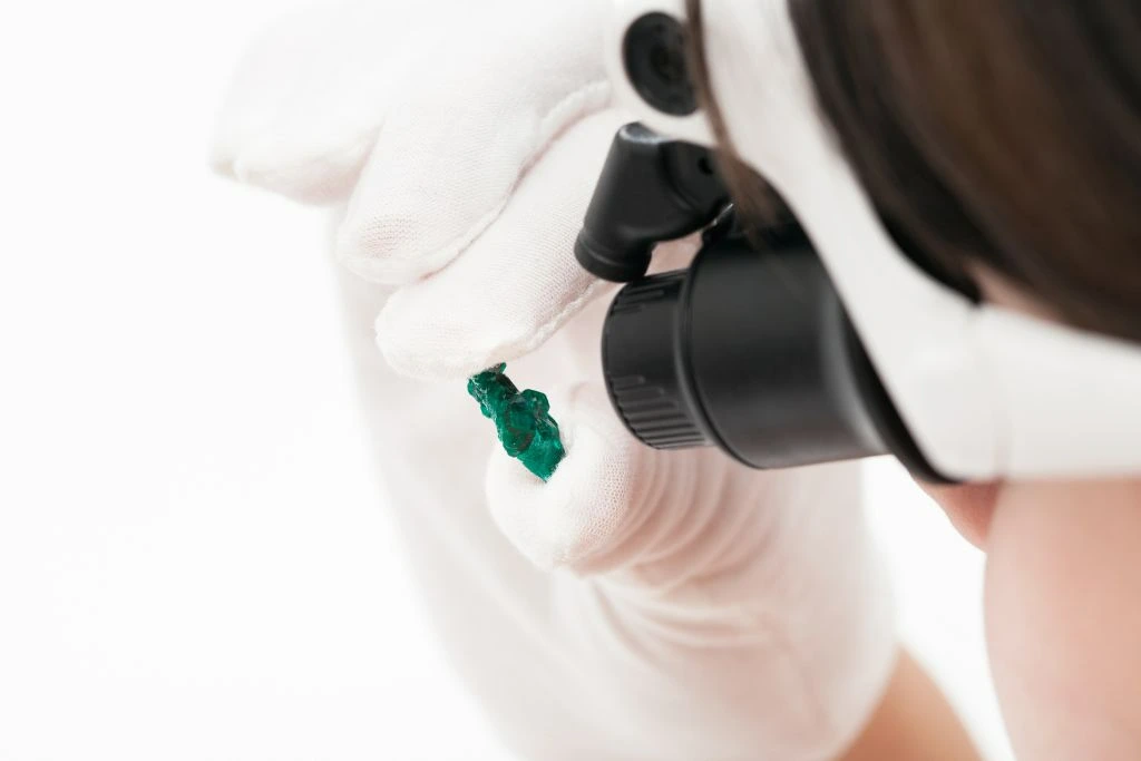 A person holding a small dioptase crystal while inspecting it using a head-mounted lens 