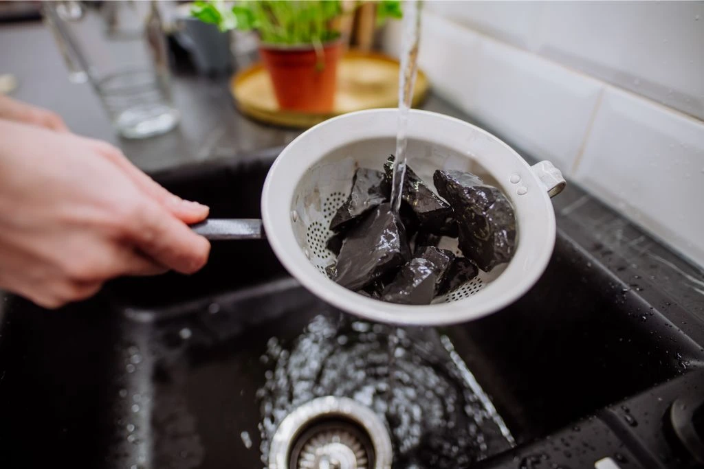 Woman Cleaning Shungite Stones in Sieve with Pouring Water in Sink