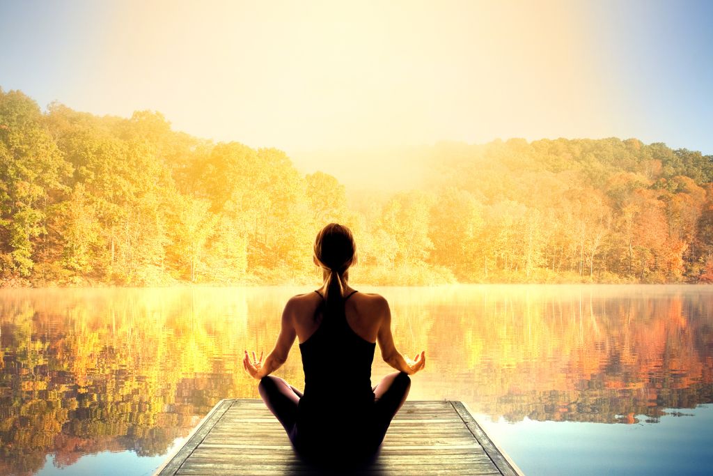 A woman is meditating near the lake while facing the sun light