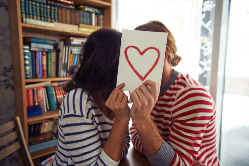 a couple covering their face behind a piece of paper with a heart written on it