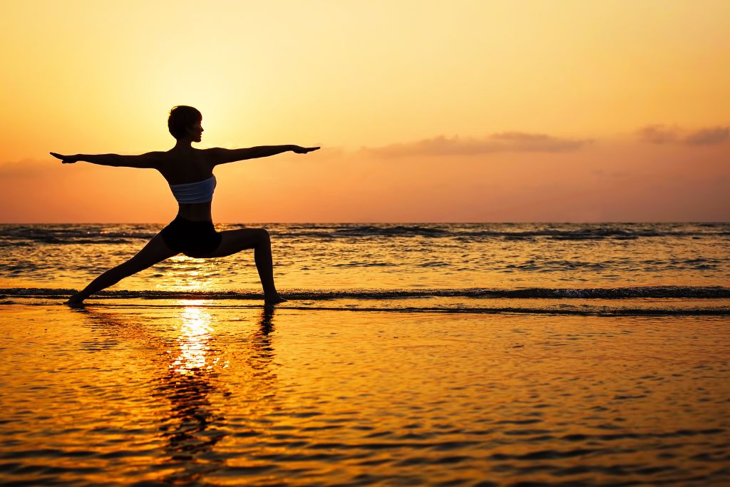 A woman is practicing yoga on the beach shore while enjoying the beautiful sunset scenery