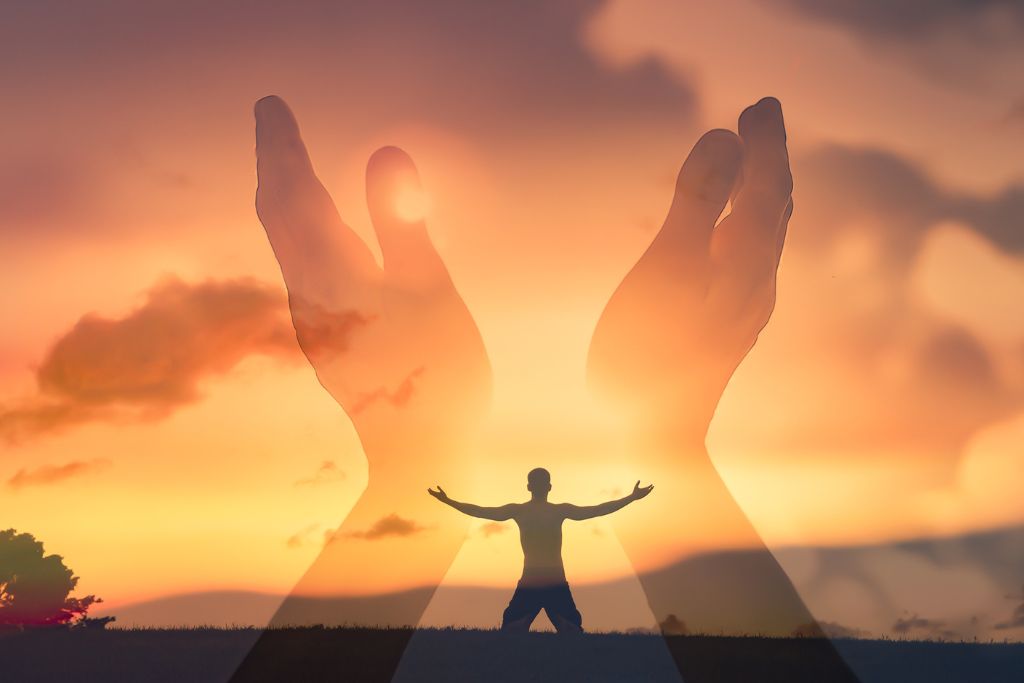 A person kneeling in an open area with the sun setting and a blurred image of a hand cupping up in the sky