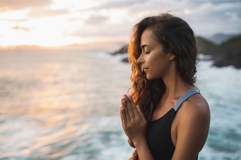 A woman praying and meditating alone at sunset with ocean and mountain