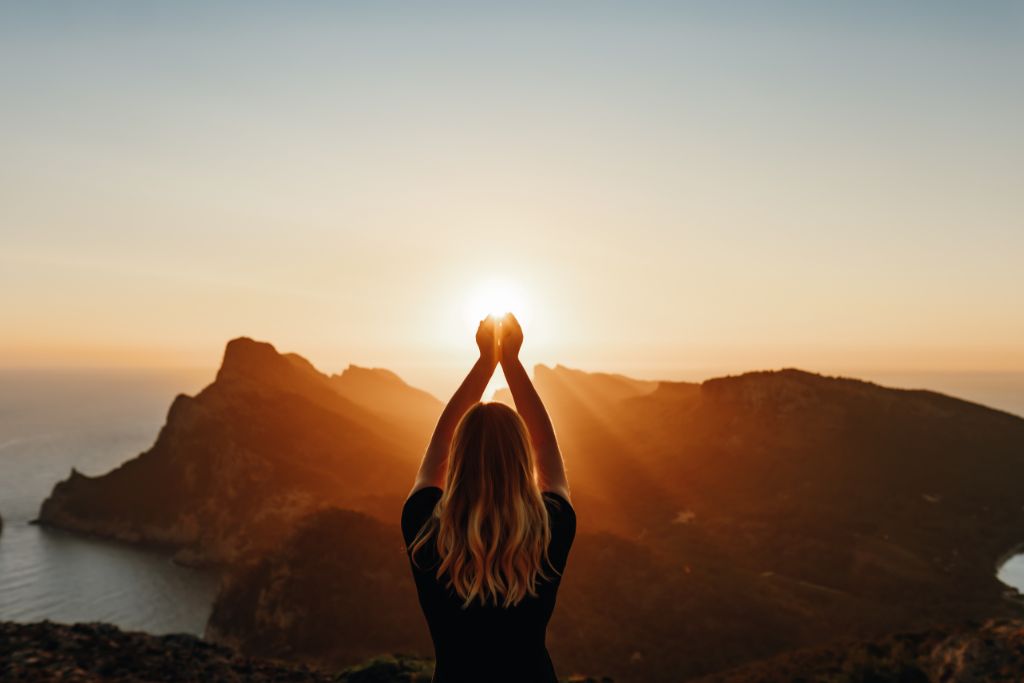 Woman meditating on top of a hill by the ocean while raising arms looking like clasping the sun. 