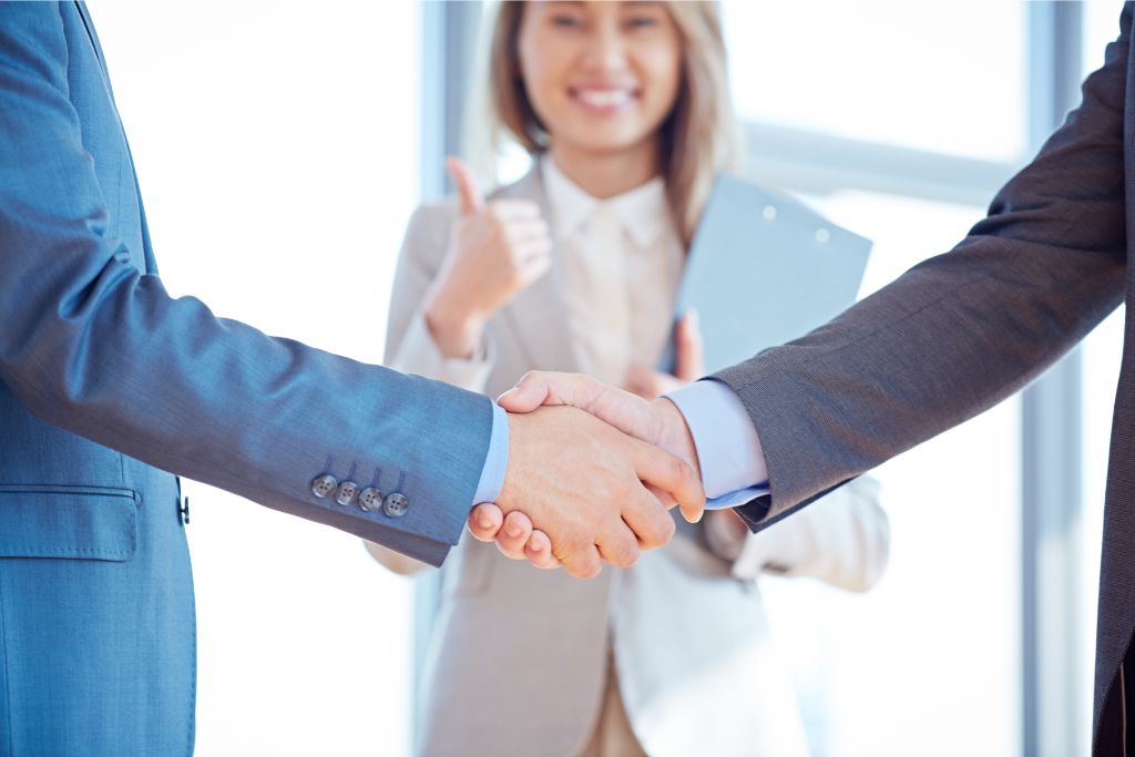 two men shaking hands and a lady in the background giving a thumbs up while smiling